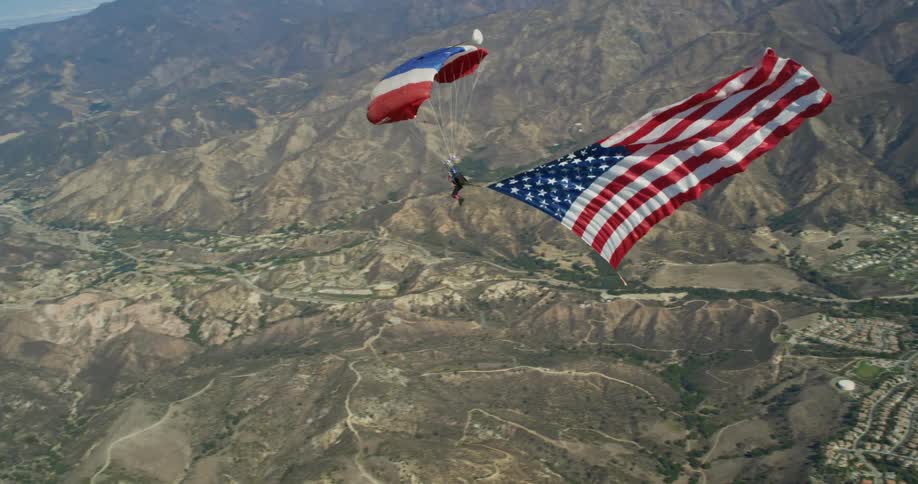 A person is flying through the sky via parachute with an american flag banner behind them