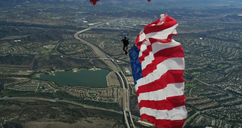 Person pulling large american flag behind them parasails over town bordered by foggy mountains