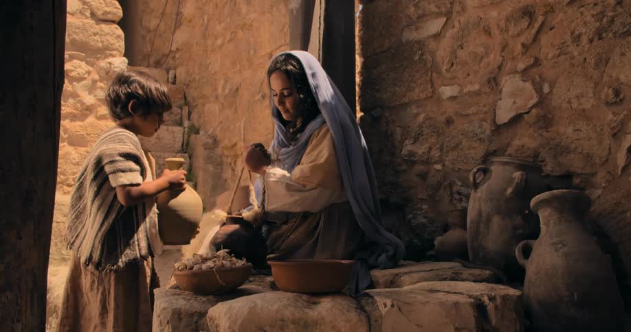 A woman sitting on a stone well pulling up a water pot from the well as a little boy walks up to her and grabs a pitcher to fill