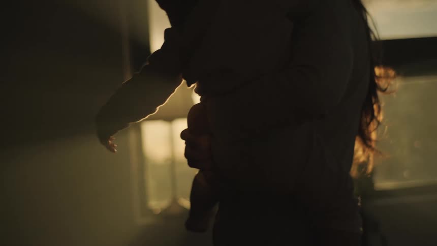 Mother holding toddler son in her harms is silhouetted agains the sun in room