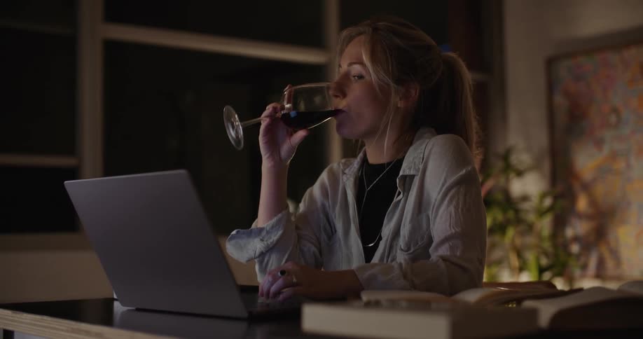 A blonde woman sips wine while working on a computer at night