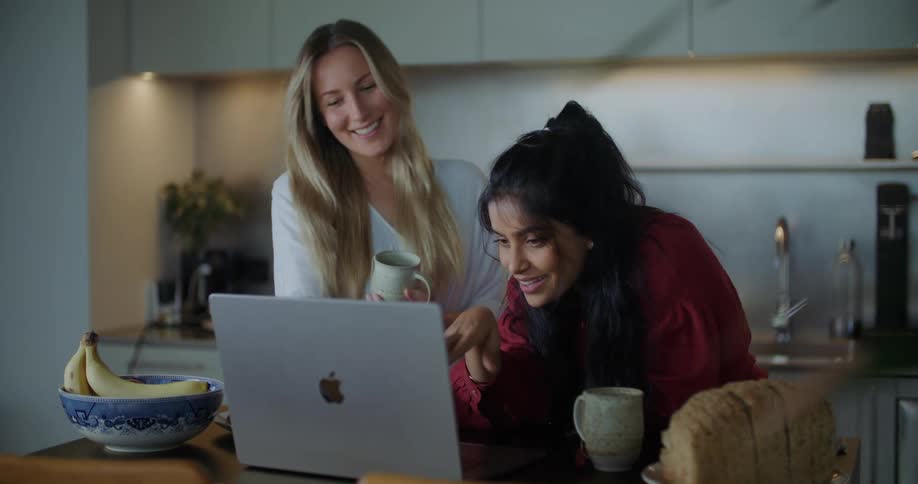 Blonde woman with white shirt and woman with red shirt look at laptop and laugh in the kitchen