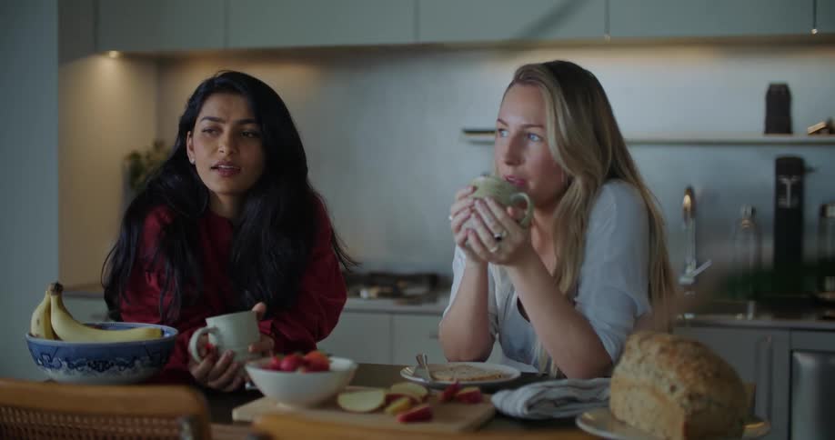 Two young women have a conversation at a kitchen table with food and tea