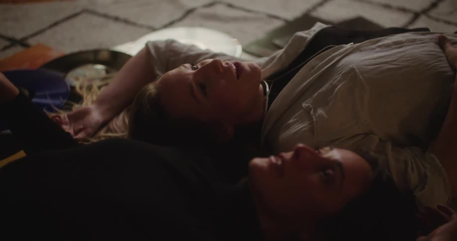 A pair of women chat with one another while lying together on a carpet with vinyl records scattered around them