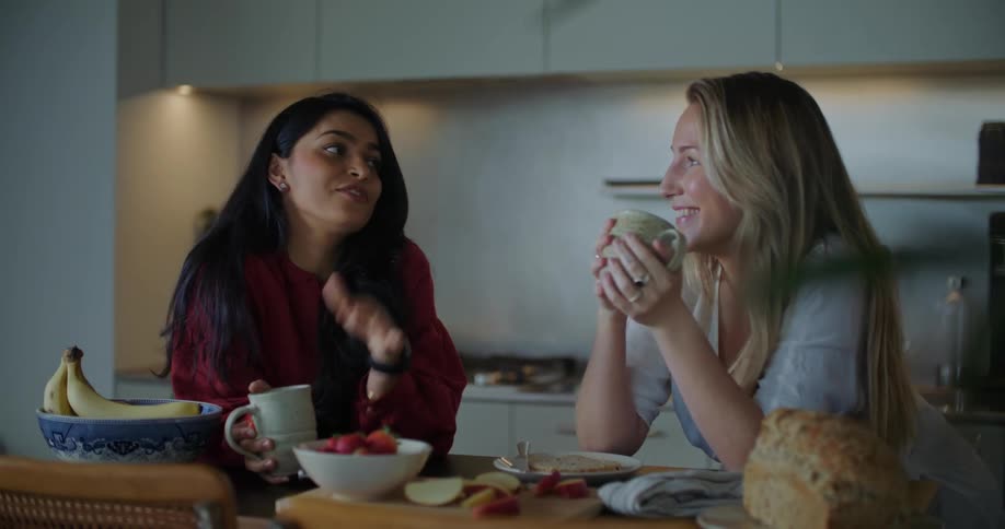Two women standing against kitchen counter chatting and having coffee and breakfast