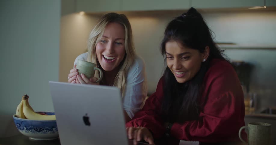 Two women are looking at a laptop computer and laughing as one woman holds a cup of coffee