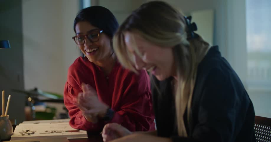 Two women smile and laugh as they talk and sit together at a table