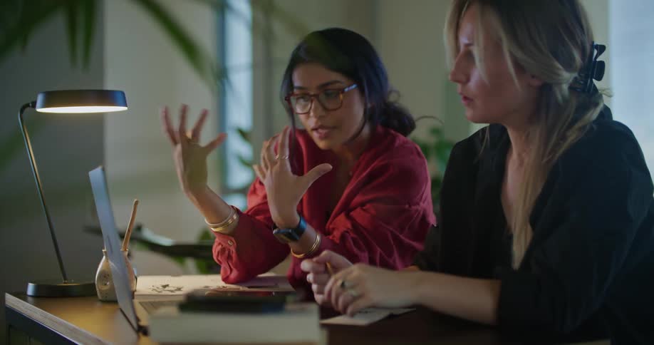 Two women are sitting at a table looking at a laptop computer and pointing to the screen as they talk and one woman is writing notes on paper
