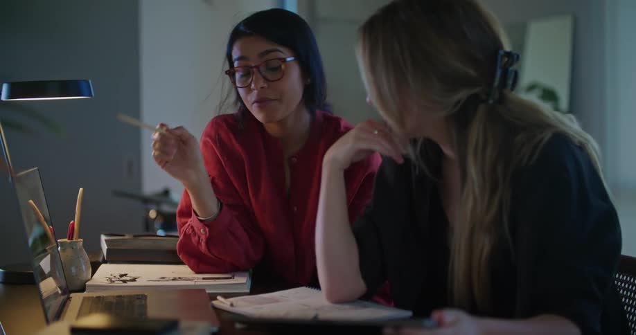 Two women are sitting at a table looking at a laptop computer and pointing to the monitor as they talk