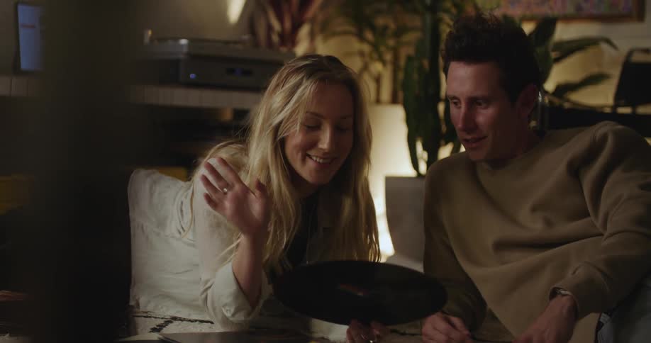 A man and a woman are sitting on the floor talking and looking at a vinyl record album