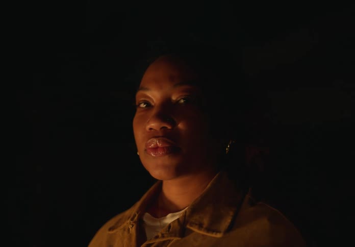 Woman with earrings and brown jacket looks at camera next to bonfire at night