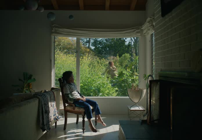 A woman walks around outside in front of a window while her son watches from inside and his brother walks over too