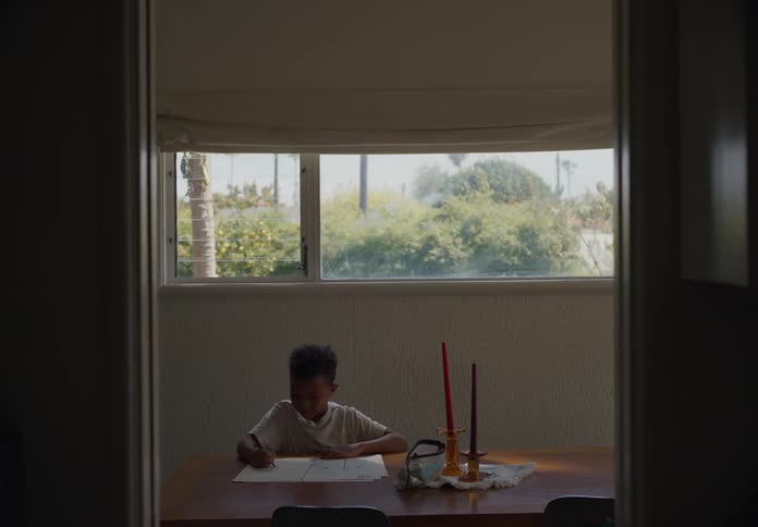 A young boy sits and draws while his brother walks over to join him