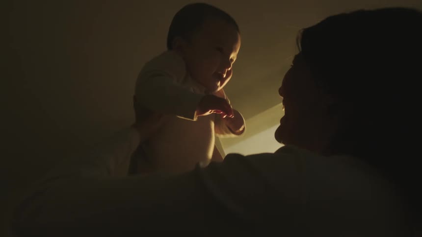 A mother stands in front of a window while playfully lifting her baby in the air