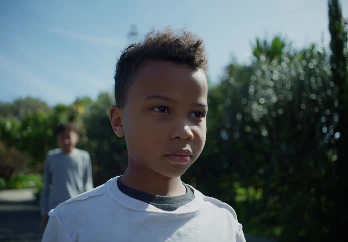 A boy standing in the road stares into the distance as an older boy comes from behind to stand beside him