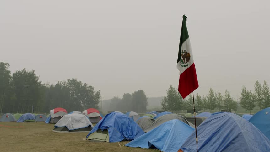 Mexican flag sits on pole near tent in full campgrounds as vehicle drives along highway on a foggy day