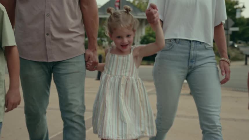 A family is walking on a sidewalk as their little girl is holding hands with both parents and smiling as she jumps
