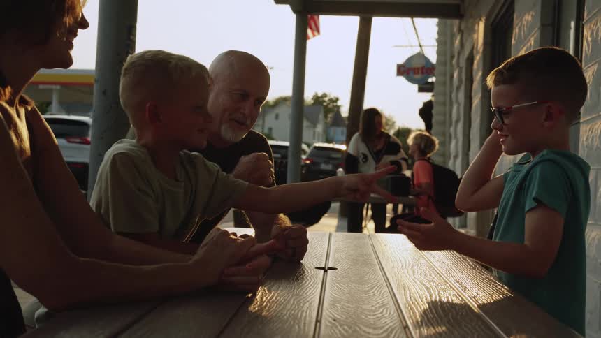Two boys sitting at an outdoor picnic table play rock paper scissors while their grandfather officiates and mother watches