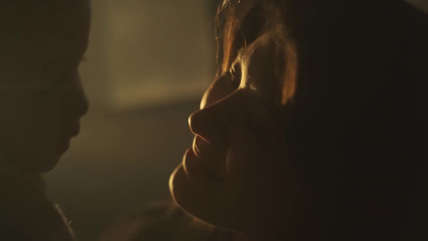 Mother with brown wavy hair holds up baby being silhouetted against the sunlight