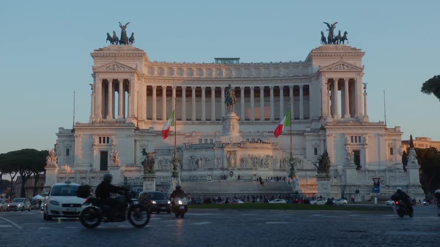 A shot of a landmark monument with national flag in the middle of a busy city square filled with passing cars and motorcycles