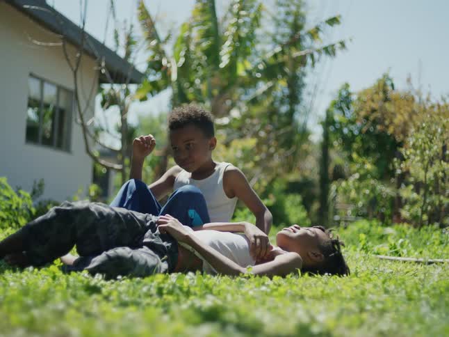 Boys with dreadlocks and white undershirts sits down and lie on green lawn in the yard