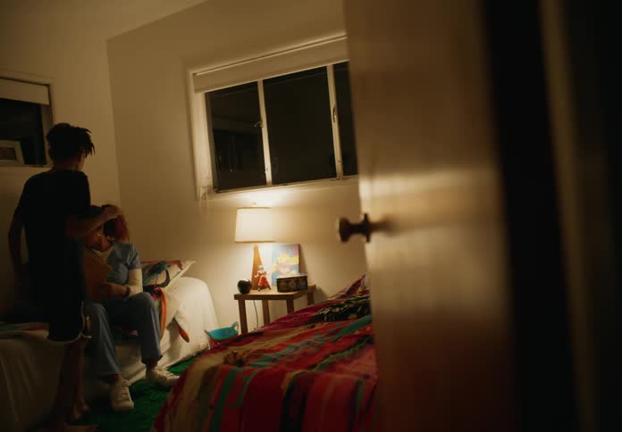 A mother sitting on her sons bed while two boys walk in and sit on the bed next to her as she opens a book