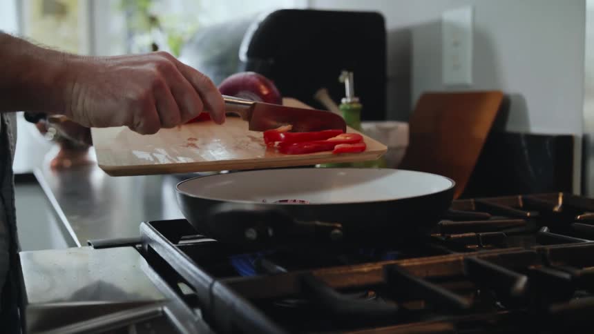 A person uses a knife to push red onions and bell peppers into a frying pan on the stove