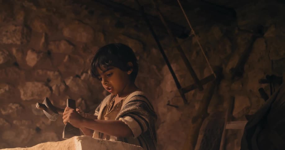 A young boy uses a hammer and chisel on a large stone under adult supervision