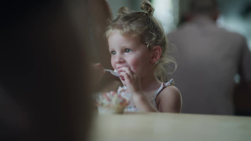 A young girl is being spoonfed ice cream from a bowl at a restaurant table