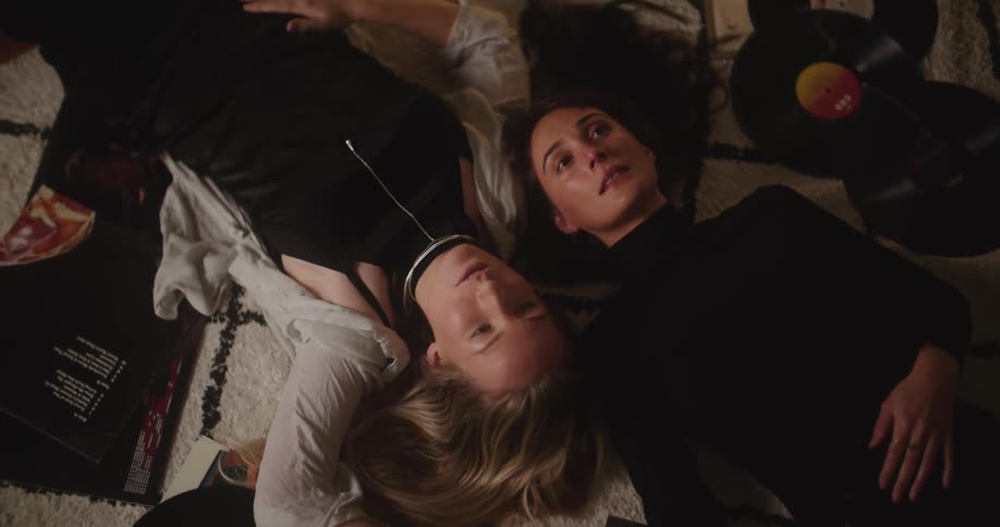 A pair of women chat while lying together on a carpet surrounded by vinyl records