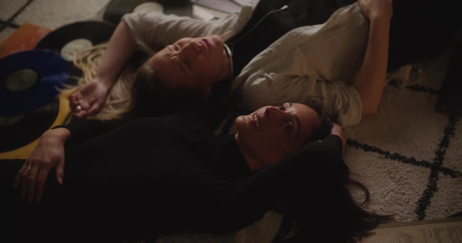Two young women are laying on the floor near some vinyl records while talking and laughing