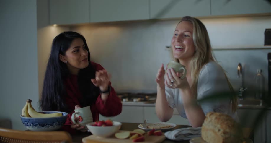 Two women standing in kitchen talking and laughing over coffee and light breakfast