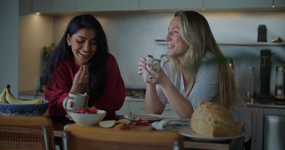Two women standing at a kitchen counter talking laughing and drinking out of mugs with food ont he coutner