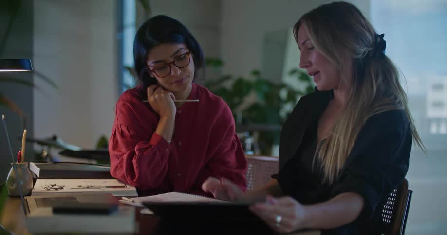 Two women collaborating on something at home at desk with laptop and papers