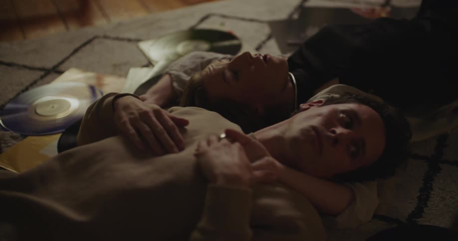 A man and woman lay on a large rug on the floor surrounded by records and holding hands while talking