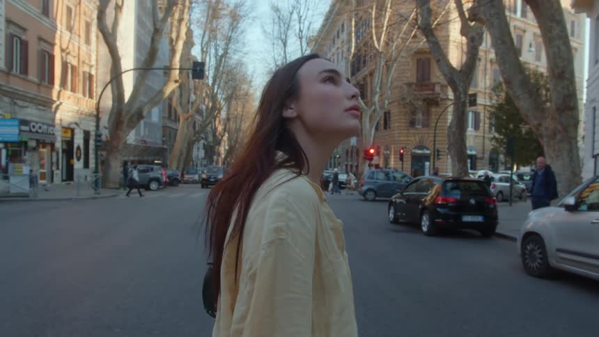 A woman is crossing a downtown street as she looks around at the buildings