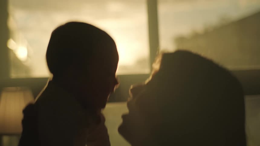 Silhouette of a mother playing with her newborn son in front of a window at evening