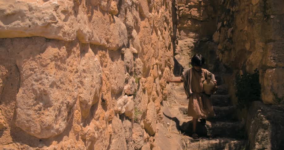 A boy in a middle eastern robe and sandals carries a vase down steps between narrow rock walls