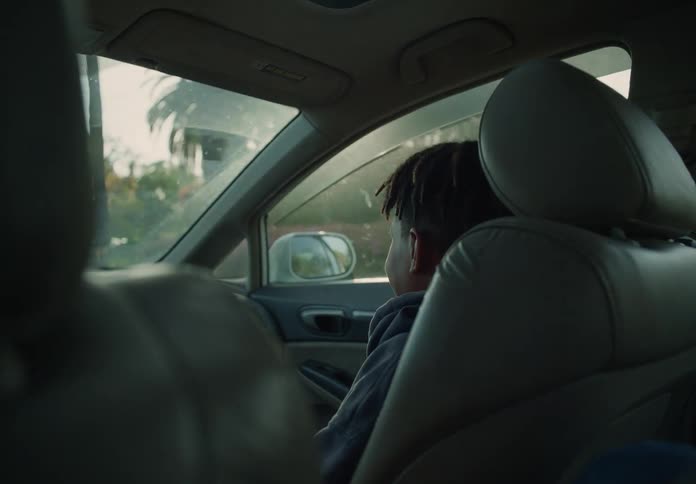 A boy sits in the passenger seat inside a moving car and smiles at the driver