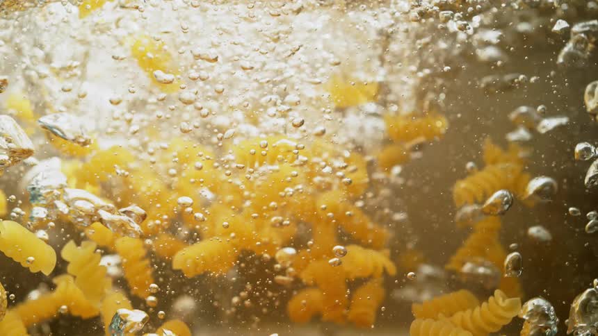 Rotini pasta noodles are dropped into water with a splash and bubbles in slow motion
