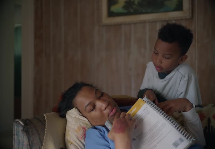 A woman is lying on her living room couch reading a book and she falls asleep as her young son walks in and covers her with a blanket