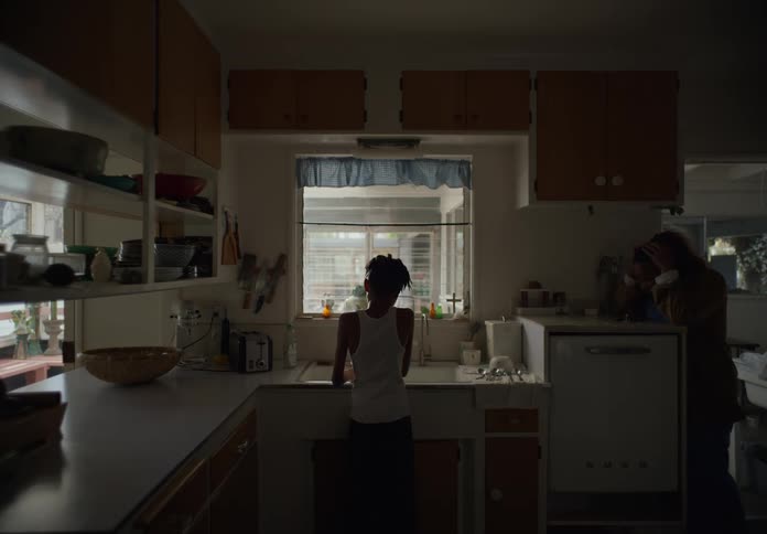 A boy standing at the kitchen sink doing dishes as his mother walks in and stands near him