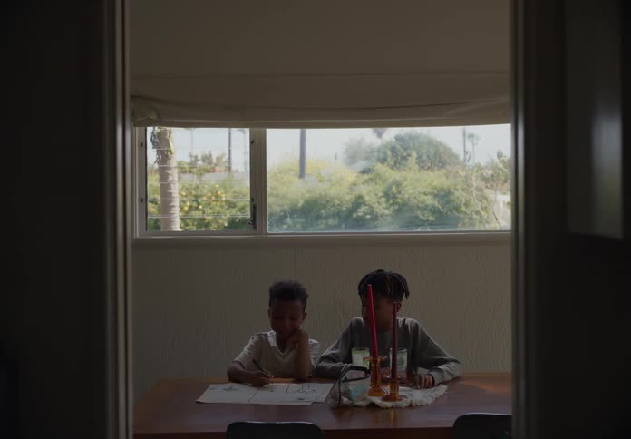 Two boys sitting at a table while one draws and the other sits and drinks from a glass