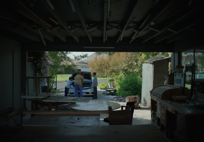 A woman leaning over the hood of a car working on it while her younger son stands next to her and her older son runs out through the garage