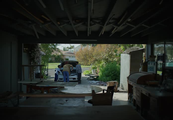 Brothers run past furniture in garage to join mother standing in front of open car trunk in driveway