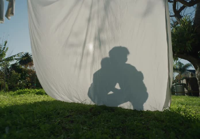 Tween brothers play fighting in silhouette behind a white sheet hanging from a clothesline in the yard