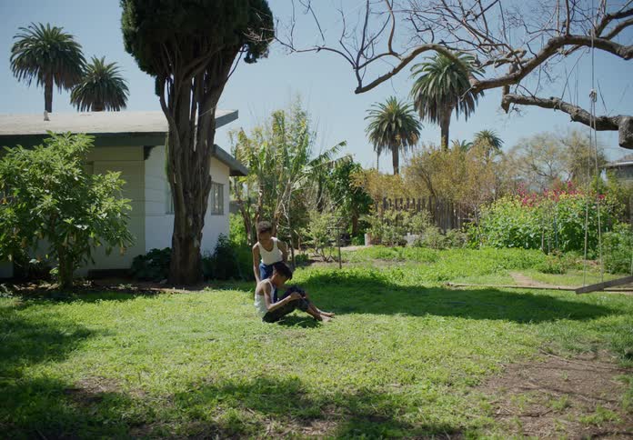 Young brothers in their backyard one laying down while the other helps him sit up and they both sit in the grass together