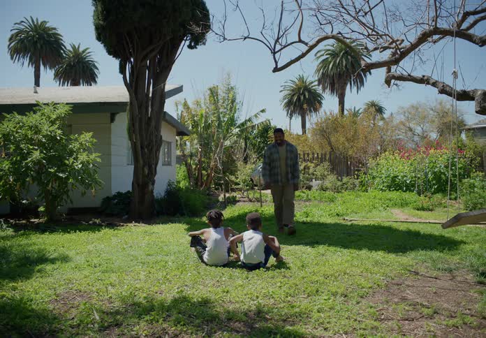 A man walks up to two young boys who are sitting on the grass in the backyard