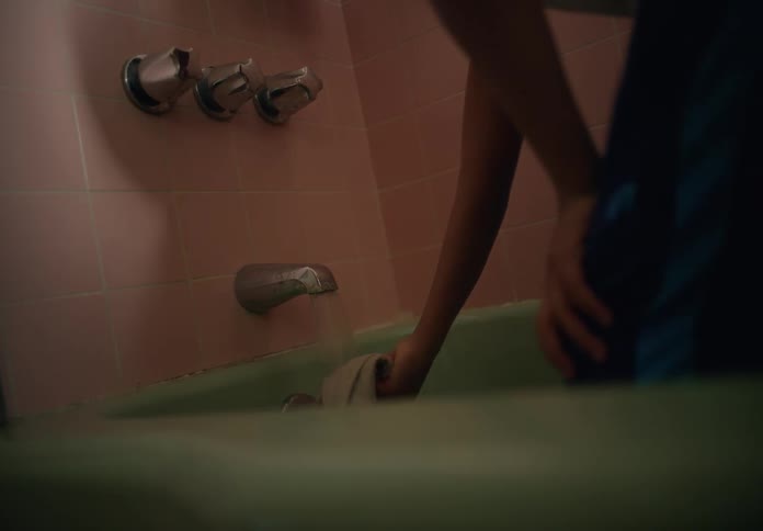 A young male steps into a bathtub turns on the faucet and completely douses a washcloth in water then turns it off