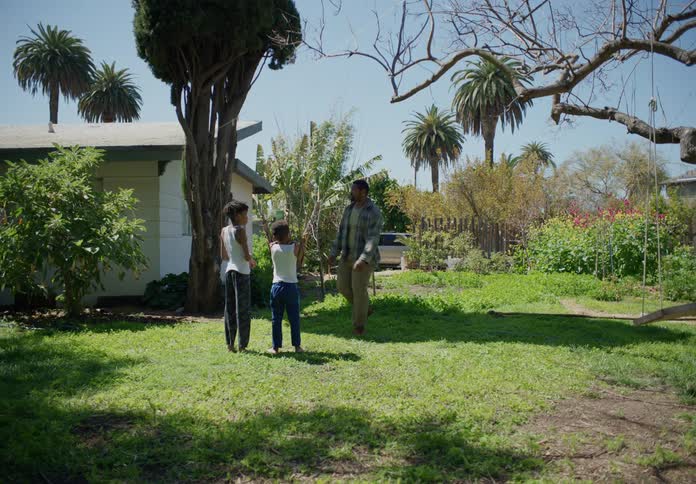 Two young boys are sitting on the grass in their yard as their father approaches
